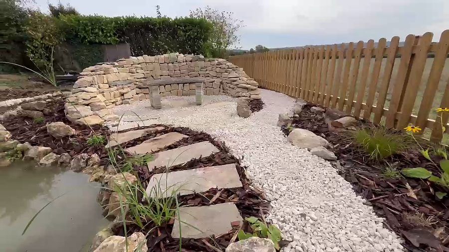Flagstone stepping stones leading past the pond to a curved drystone wall with a seat built into it